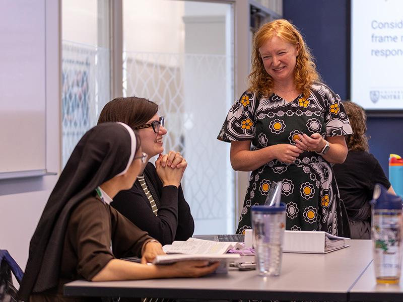 Professor stands next to two students at their desks, studying Catholic leadership